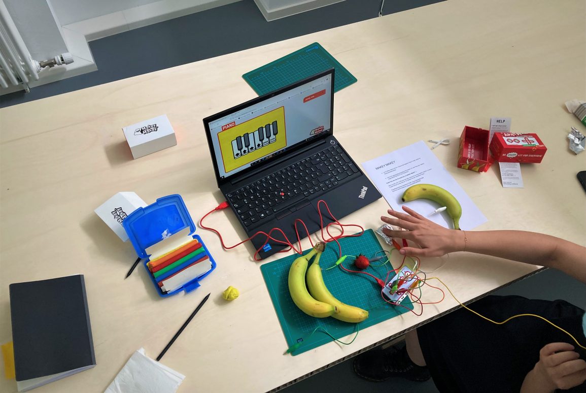 Von oben sehen wir einen Laptop, an den mit rotem Kabel eineMakey-Makey-Leiterplatte angeschlossen ist. Aus dem Makey Makey gehen viele Kabel raus. Teilweise sind die Kabel an die Objekte auf dem Tisch angeschlossen. Dazu zählen die Bananen, die Erdbeere und etwas gefaltete Alufolie. Ein Kabel wird von einer Person in der Hand gehalten, während mit der anderen Hand die Alufolie berührt wird. Ein grünes Kabel ist nirgends angeschlossen. Auf dem Bildschirm des Laptops wird eine Klaviertastatur angezeigt. Auf dem Tisch verteilt liegen zusätzlich noch bunte Knetestifte in einer blauen Box, zur Kugel gerollte gelbe Knete, Bleistifte, ein Notizbuch, Bastelmatten, die Anleitung zum Kennenlernen des Makey Makeys sowie dessen Verpackung.