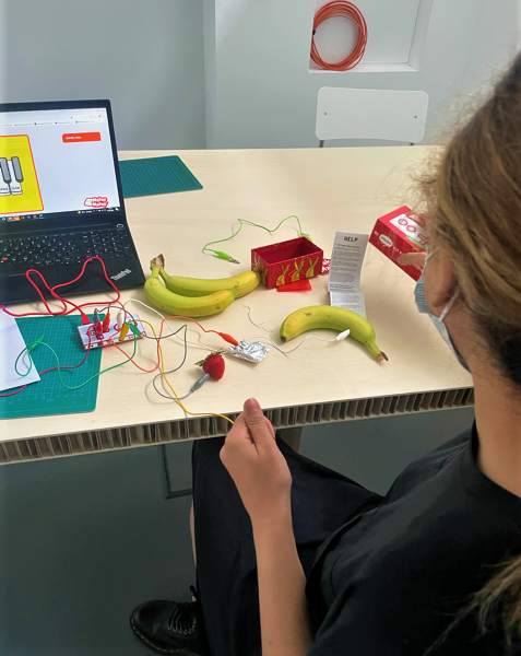 Nirvana hält ein Kabel in der linken Hand. Dieses ist mit dem Makey Makey verbunden. Vom Makey Makey aus führen weitere Kabel zu gefalteter Alufolie, einer Banane und einer Erdbeere. Dahinter steht ein Laptop auf dem Tisch. Auf dem Bildschirm ist der Ausschnitt einer Klaviertastatur zu erkennen. Auf dem Tisch liegen weitere Bastelmaterialien verstreut.