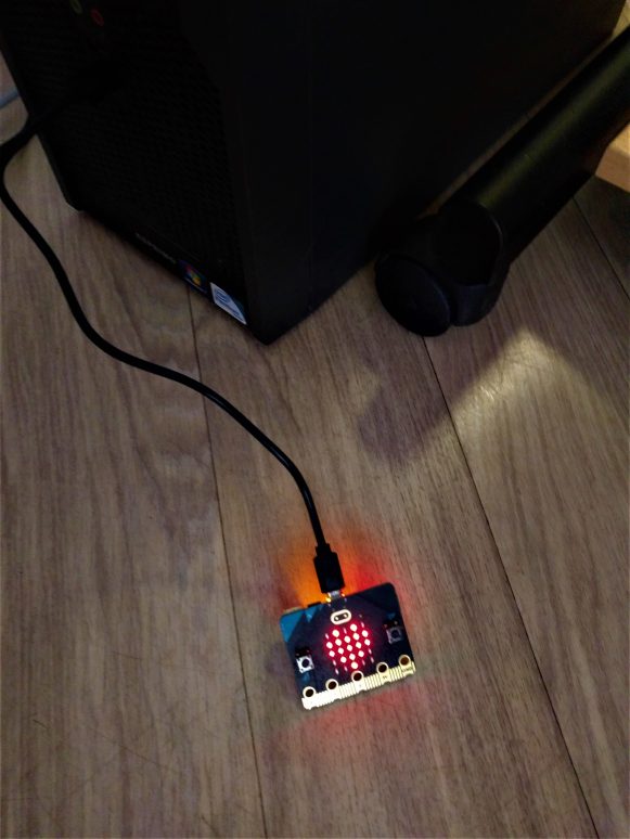 Ein auf dem Boden liegender micro:bit zeigt ein leuchtendes, rotes Herz auf der 5×5-LED-Matrix. Der micro:bit ist per USB an einen Rechner angeschlossen.