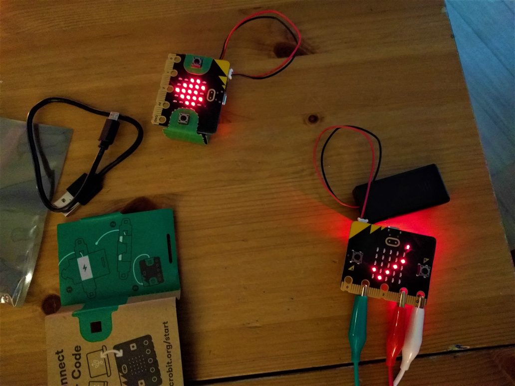 Auf dem Tisch liegen zwei micro:bits, die über Batterien mit Strom versorgt werden. Ein micro:bit ist direkt auf den Batteriehalter montiert und zeigt ein großes Herz auf der 5×5-LED-Matrix. Das andere micro:bit liegt neben dem Batteriehalter und zeigt einen Haken. Zudem hört es auf
Umgebungsgeräusche, da die LED für das Mikrofon leuchtet. Unten an
den Pins sind drei Kabel per Krokodilklemme angeschlossen: Grün an Pin1 (Daten), Rot an 3V (Stromversorgung) und Weiß an GND (Masse).